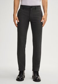 Mann in slim-fit, dunkelgrauen karierter Hose und schwarzen Leder-Schnürstiefeln, stehend vor einem neutralen Hintergrund.