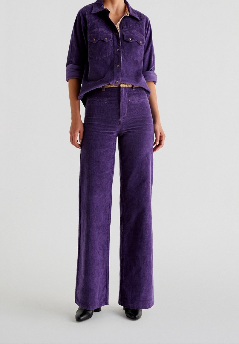 Femme portant une chemise en velours côtelé violette à boutons avec manches retroussées et un pantalon large taille haute assorti, associée à des chaussures à talons noires.