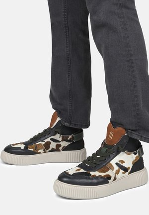 Baskets montantes avec un textile imprimé vache, des accents en cuir noir, une semelle en caoutchouc texturée, et des détails en vert olive sur les lacets et le talon.