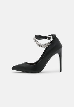 Schwarzer Leder-Pump mit hohem Absatz und spitz zulaufender Zehenpartie, ausgestattet mit einem silberfarbenen Kettenknöchelriemen und einer eleganten, strukturierten Oberfläche.