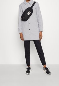 Vestido camisa gris claro de manga larga con botones; riñonera negra con logo; pantalones negros ajustados; zapatillas negras con detalles blancos.