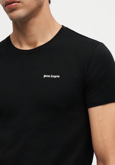 Άνδρας που φοράει μαύρο κοντομάνικο T-shirt με λευκό κείμενο "Palm Angels" στο αριστερό στήθος, σε απλό φόντο.