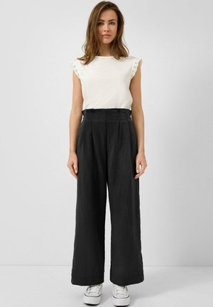 Junge Frau steht frontal und trägt ein weißes ärmelloses Oberteil mit Spitzenbesatz, hoch taillierte, weit geschnittene schwarze Hosen und weiße Sneakers.