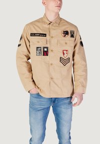 Beige U.S. Army jakke med broderede mærker, knaplukninger, to forlommer og en afslappet pasform. Udstyret med forskellige insignier.