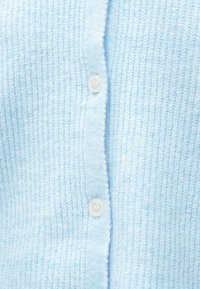 Svetlo modra pletenina z dvema prosojnima belima gumboma, zapetima v navpični vrsti na cardiganu.