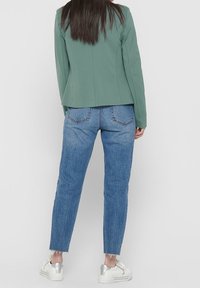 Grön blazer med strukturerade axlar, kombinerad med ljusblå jeans med fransad fåll och silverfärgade sneakers, sedd bakifrån.