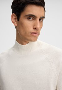 Maglione color crema con collo alto e bordo a coste. Tessuto a maglia con maniche raglan. Design aderente che mette in risalto la parte superiore del corpo.