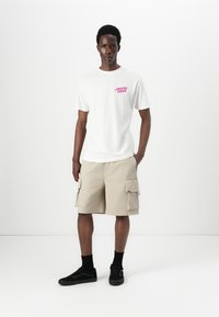 White cotton t-shirt with pink graphic text, paired with beige cargo shorts featuring side pockets. Worn with black sneakers and socks.
