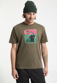T-shirt en coton vert olive avec une illustration colorée d'une figure entourée d'un soleil et d'un arbre. Manches courtes et col rond.