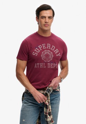 Mężczyzna ubrany w bordowy T-shirt z napisem "Superdry Athl Dept", niebieskie jeansy i koszulę w kratę zawiązaną wokół talii, stoi na białym tle.