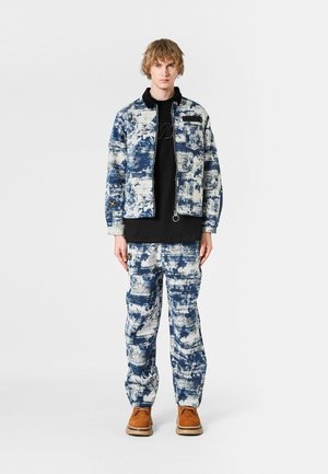 Chaqueta de patrón azul y blanco con cierre de cremallera, combinada con pantalones a juego; ambos confeccionados en tejido texturizado. Zapatos marrones complementan el conjunto.