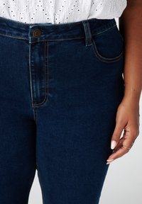 Hoge taille donkerblauwe denim jeans met een gladde textuur, zichtbare stiksels en zijzakken; een metalen knoop aan de taille.