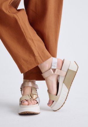 Pies con sandalias de plataforma beige y metálicas con suelas gruesas, combinadas con pantalones de color óxido sobre un fondo liso y claro.