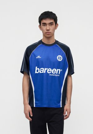Junger Mann trägt ein blau-schwarzes Sportshirt mit weißem Schriftzug "bareen motorsport" und steht vor einem einfarbigen hellen Hintergrund.