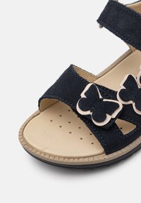 Marinblå mockasandal med två justerbara remmar, beige vadderad fotbädd och rosa fjärilsdetaljer. Flexibel gummisula med perforeringar.