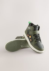 Sneakers alte in materiale sintetico verde oliva con accenti neri, dotate di una fascia in Velcro, un anello verde brillante e patch giocosi.