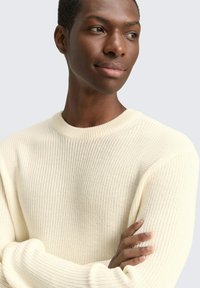 Crème gebreide trui met ronde hals en ribgebreide manchetten, met een gestructureerde weving. Model poseert met gekruiste armen, tegen een neutrale achtergrond.