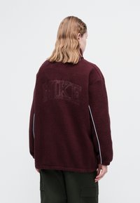 Burgunderfleecegenser med høy krage, med en preget logo på baksiden og kontrastpiping langs ermene. Teksturlignende stoff.