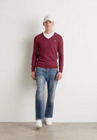 Pulover bordo cu decolteu în V peste o cămașă albă, asortat cu blugi denim deschiși la culoare și adidași albi. Modelul poartă o șapcă bej.