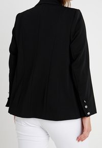 Blazer nero con tessuto testurizzato, vestibilità strutturata e colletto curvo. Presenta tre dettagli in bottoni argentati sulle maniche.