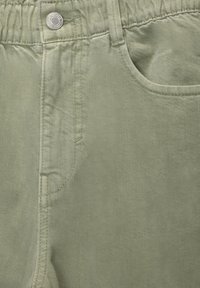 Primer plano de pantalones de algodón verde claro que muestra la cinturilla elástica, el botón de metal, el bolsillo delantero y las costuras detalladas.