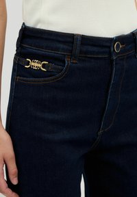 Mörkblå denimjeans med guldfärgad hårdvara på framfickan och orange sömmar längs sömmarna. Fitted design med bältesömmar.