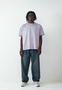 Jasnofioletowa bawełniana koszulka z krótkim rękawem, zestawiona z luźnymi niebieskimi dżinsami i szarymi trampkami. Widoczny detal w postaci prostej łańcuszkowej kolii.