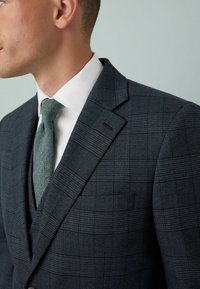 Jachetă de costum în carouri bleumarin cu un finisaj texturat. Dispune de rever clasic, un buzunar și o cravată verde asortată.
