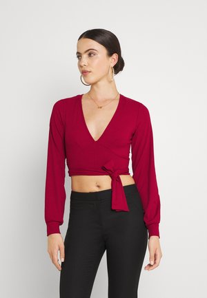 Top cropped de mangas longas em malha vermelha com decote em V profundo e detalhe de laço frontal, combinado com calças pretas de cintura alta.