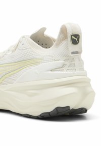 Zapatilla de correr ligera con un upper texturizado en color crema, rayas de acento amarillas y una suela blanca gruesa. Presenta un logo de Puma prominente en el talón.