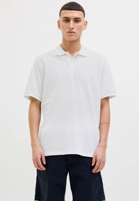 Polo shirt bianco a maniche corte con colletto classico, texture liscia, vestibilità rilassata, indossato con pantaloni corti scuri, con un sottile dettaglio del logo sul lato.