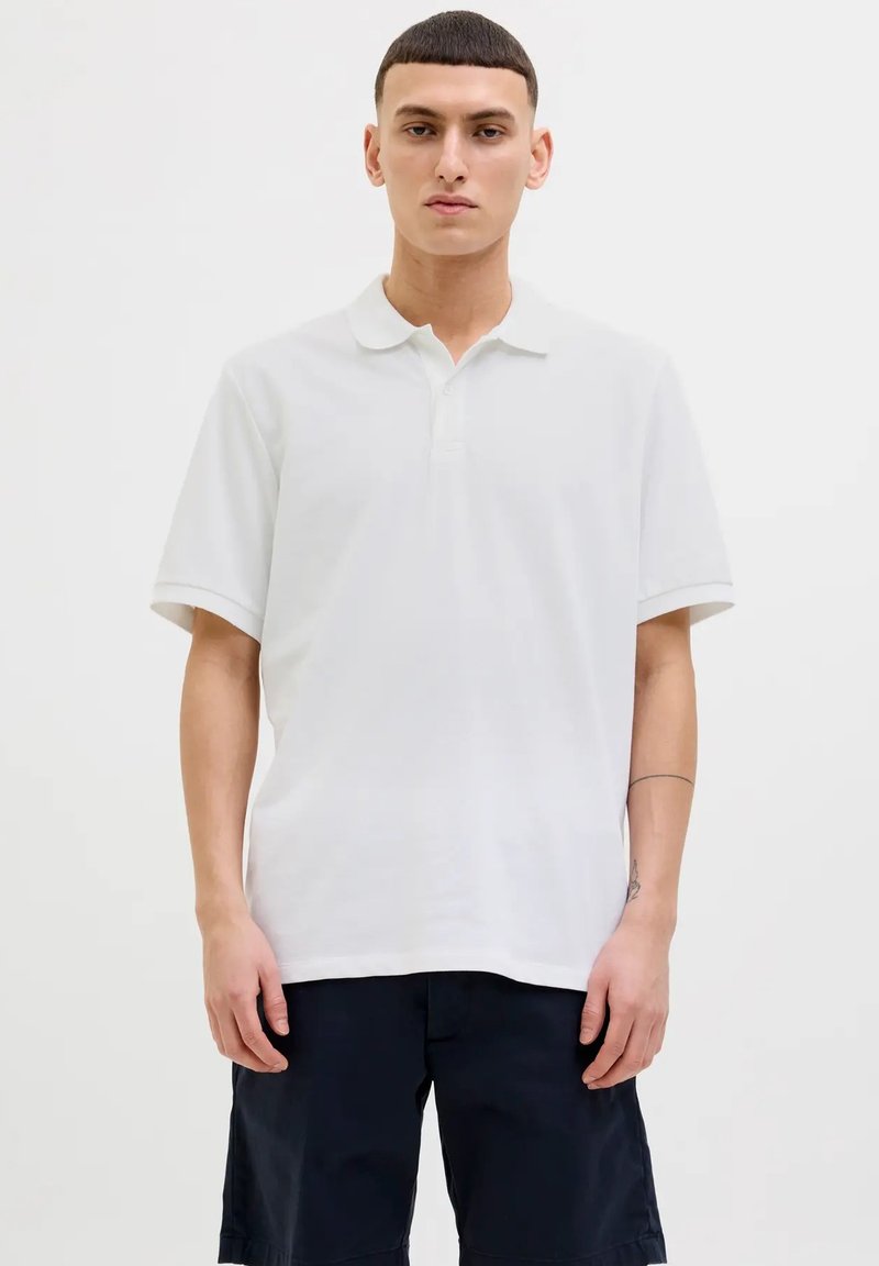 Polo shirt bianco a maniche corte con colletto classico, texture liscia, vestibilità rilassata, indossato con pantaloni corti scuri, con un sottile dettaglio del logo sul lato.