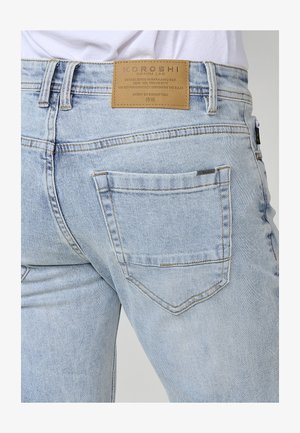 Világoskék farmernadrág magas derekkal, hátul bőr címkével, amelyen a "KOROSHI" felirat szerepel, valamint jellegzetes hátsó zsebfedések varrásával.