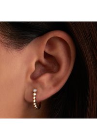 Boucle d'oreille en or avec une rangée de petites perles et un accent en diamant, visible à côté d'une oreille avec des cheveux foncés. Texture lisse et forme ronde.
