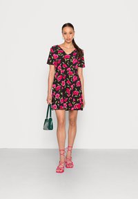 Vestido preto floral com flores cor-de-rosa, mangas curtas, decote em V e cintura ajustada. Combinado com sandálias cor-de-rosa com tiras e uma bolsa verde.