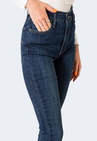 Vaqueros de tiro alto de color azul oscuro en material de mezclilla con una textura suave; cuenta con cinco bolsillos y acentos de costura visibles.