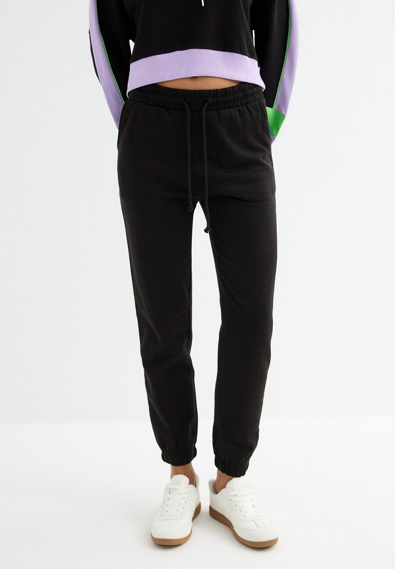 Pantalones jogger negros con cintura y puños elásticos, fabricados en suave tela de algodón. Combinado con zapatillas blancas para un look casual.