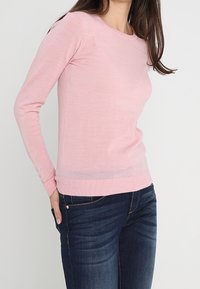 Pull en tricot rose pâle avec un col rond, des manches longues et des poignets côtelés. Associé à un jean bleu foncé. Texture lisse.