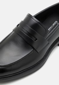 Mocasines de cuero negro con suela plana. Presenta una correa decorativa en la parte superior y costuras visibles a lo largo del borde. Textura suave.