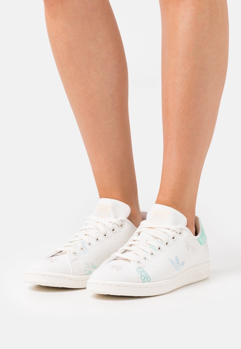 Baskets basses blanches Adidas avec des détails bleu clair portées pieds nus, présentées sur un fond blanc uni.