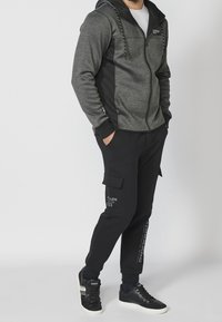 Sweat à capuche zippé gris et noir avec un col montant, associé à un pantalon de jogging noir avec des poches cargo. Les deux sont fabriqués en tissu doux et texturé.