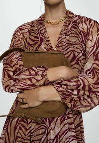 Femme portant une robe à imprimé zèbre violet et beige, tenant un sac à main en daim marron contre sa poitrine, avec un collier en chaîne dorée.