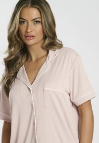 Ljusrosa kortärmad pyjamasöverdel med ribbad textur, vit piping runt kragen och en enda framficka. Knäppning med knappar.