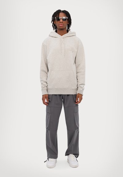 Haut à capuche gris clair, coupe décontractée avec une poche avant, associé à un pantalon cargo gris foncé. Des baskets blanches et des lunettes de soleil noires complètent le look.