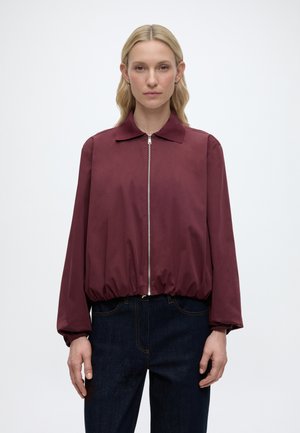 Blonde Frau mit glattem Haar trägt eine bordeauxrote Reißverschlussjacke mit elastischem Saum und dunkelblaue Jeans, stehend vor einem einfarbigen weißen Hintergrund.