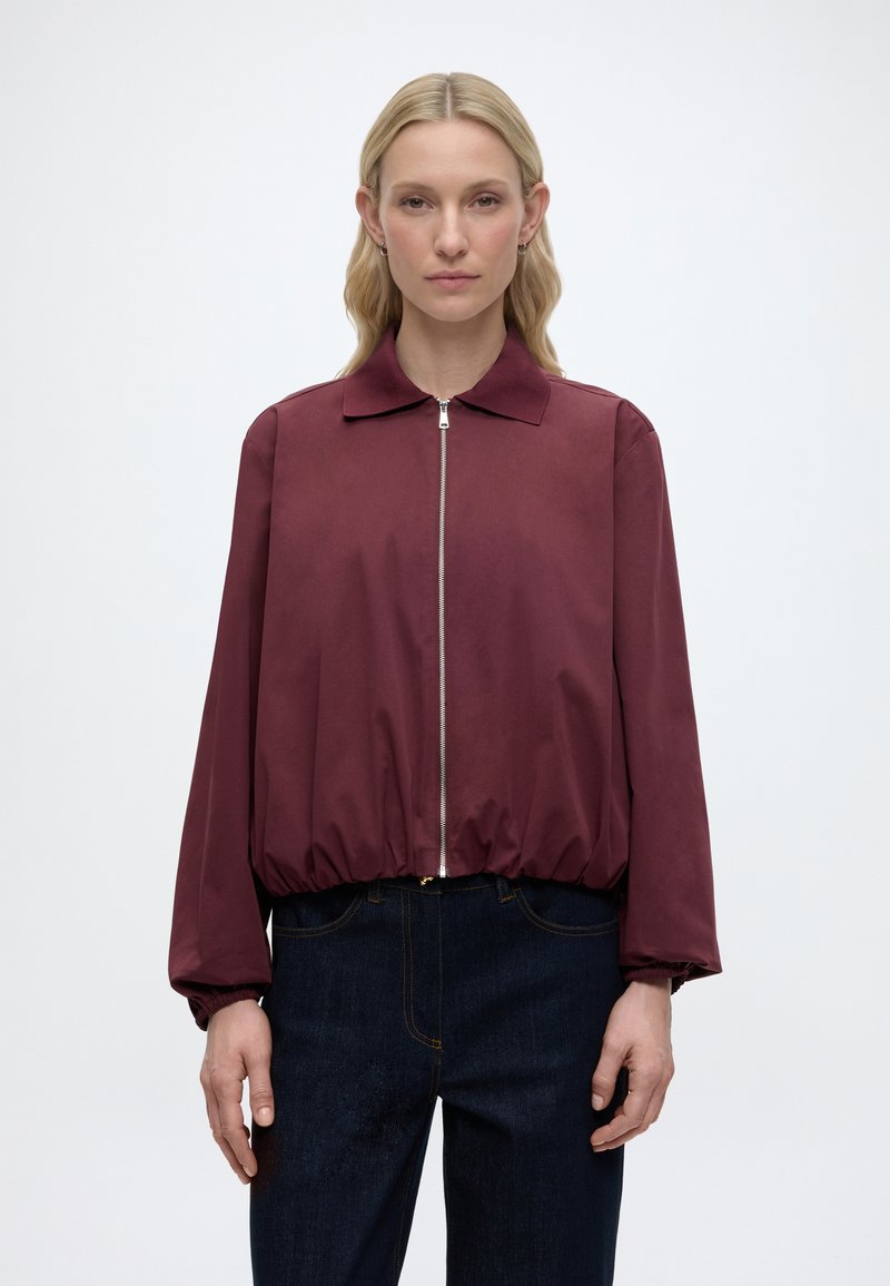 Sirgete sirged juustega blond naine kannab burgundi värvi lukuga jope elastse ääre ja tumesiniste teksadega, seisab ühevärvilise valge tausta ees.