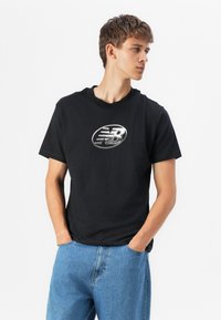 T-shirt en coton noir avec encolure ronde et un logo ovale argenté sur la poitrine. Associé à un jean en denim bleu clair. Texture lisse.