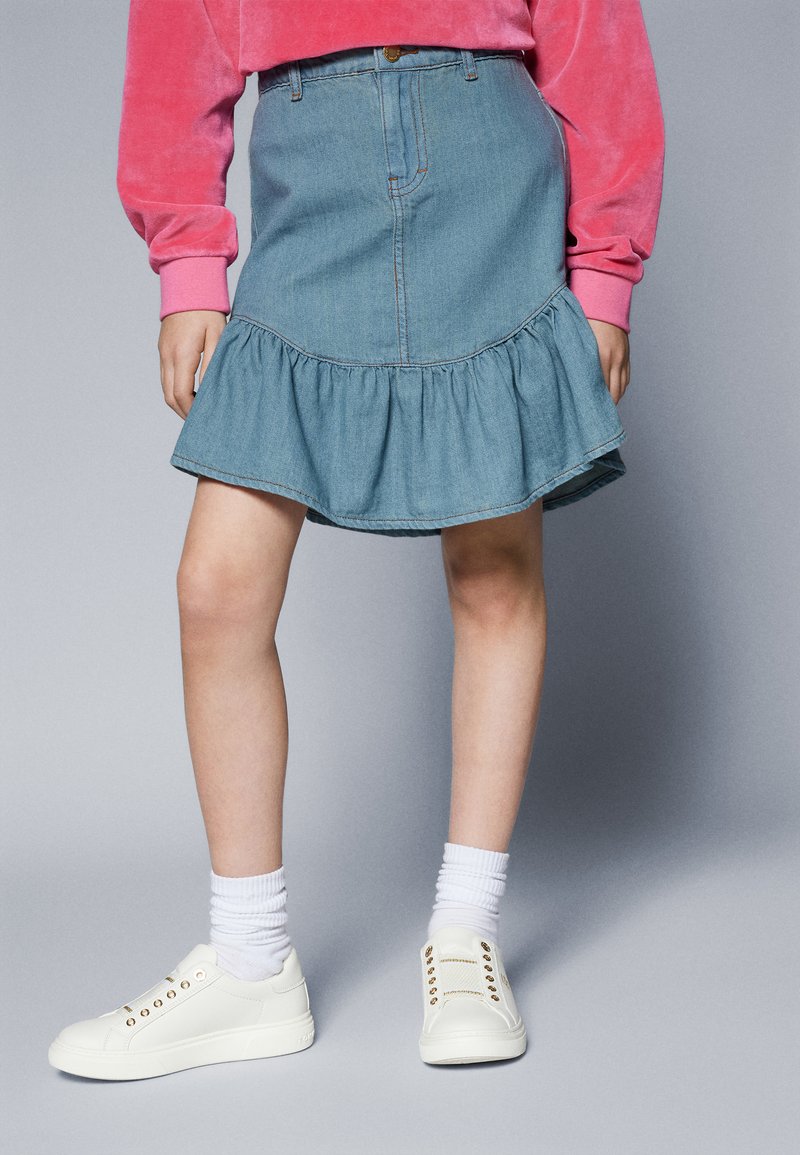Jeanskjørt i lys blå med volangkant, kombinert med hvite joggesko og ribbestrikkede hvite sokker. Glatt tekstur og avslappet design.