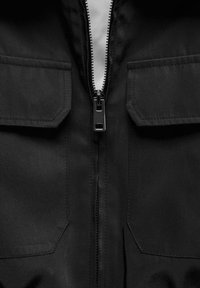 Veste noire avec une fermeture éclair centrale, deux poches poitrine, et un tissu durable à texture lisse. Conçue avec des rabats de poche angulaires.