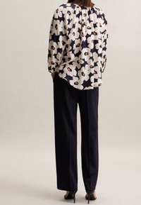 Blusa fluida con motivo floreale scuro nei colori crema e corallo, abbinata a pantaloni neri a vita alta e tacchi neri.